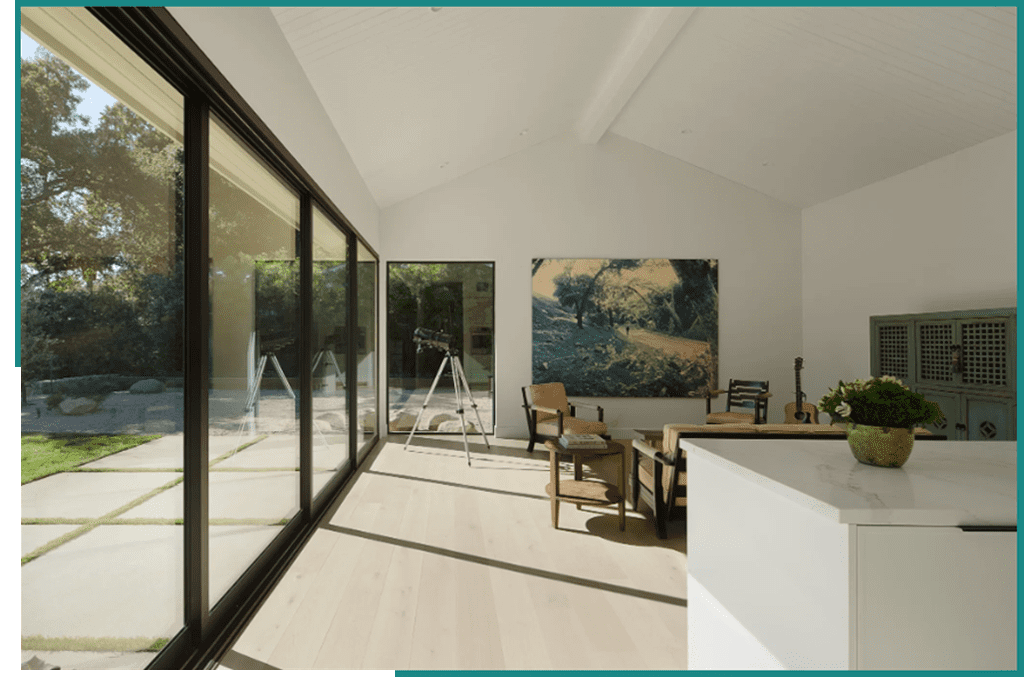 inspired-2 A spacious, modern room with a high white ceiling, large glass windows showcasing an outdoor view, wooden flooring, a simple chair with a footrest by an easel, artwork on the wall, and.