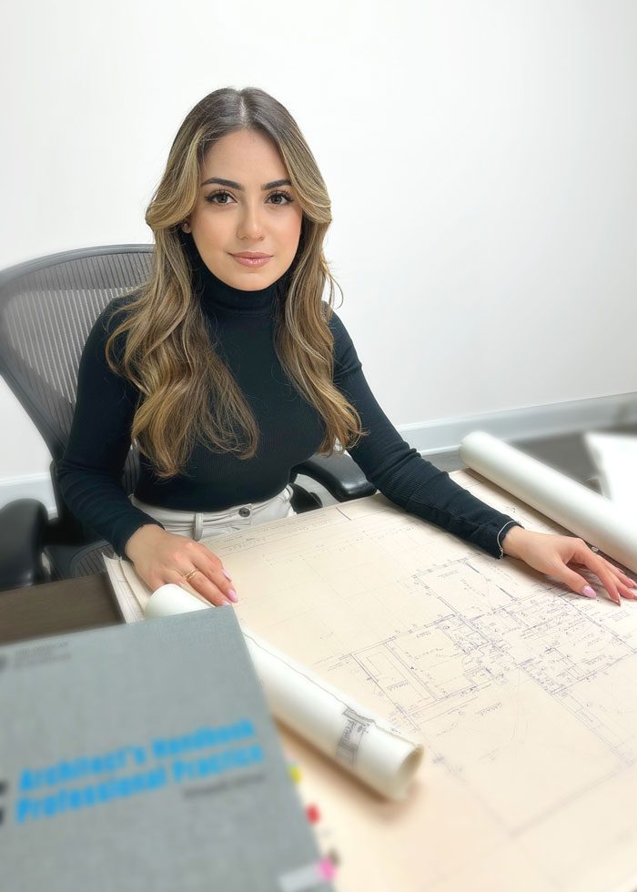 Kristine-01-Color-Final A woman in a black turtleneck sitting at a desk with blueprints and an engineering textbook, looking at the camera.