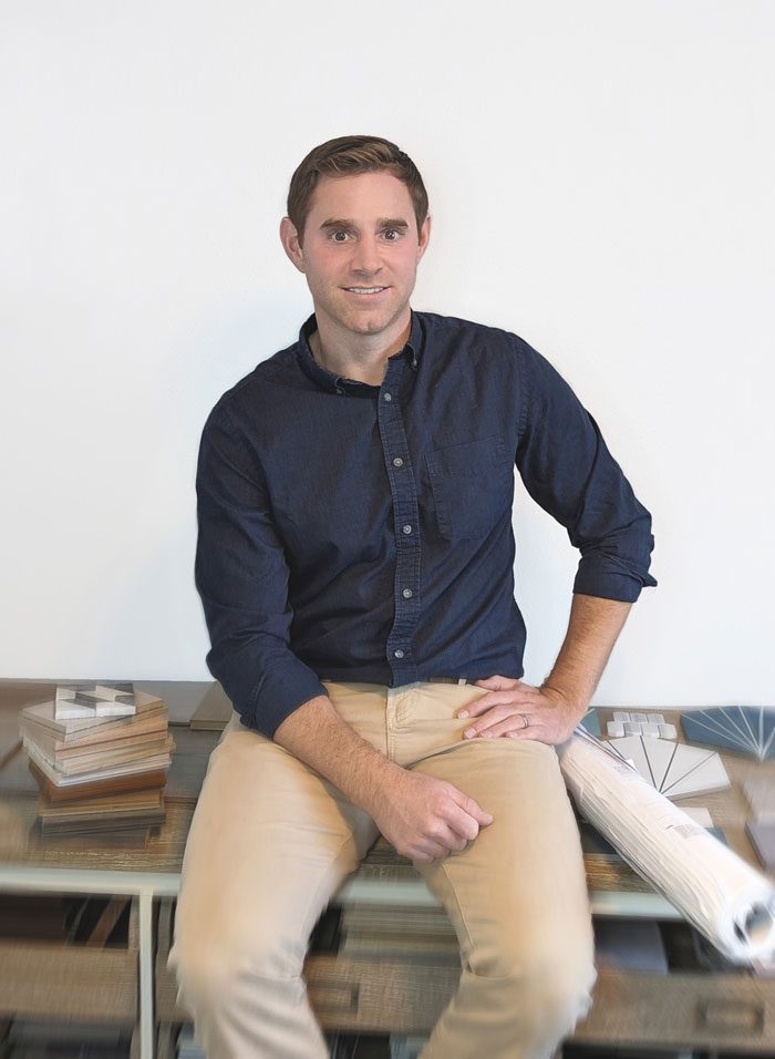 MarkNichols2023Headshot-Final-withBlur Man in a dark blue shirt and beige pants sitting on a desk, smiling at the camera, with books and plans beside him.