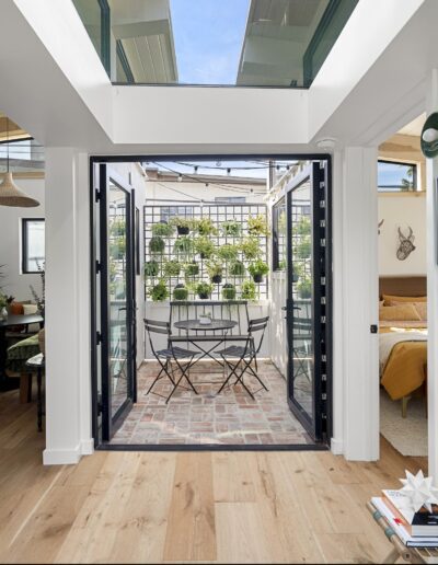 Compact indoor terrace with brick flooring, black metal-framed glass doors, and several small potted plants hanging on a grid. Adjacent rooms visible include a dining area and a bedroom.